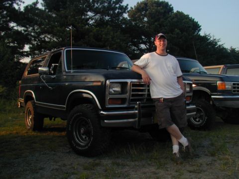 1988 Ford Bronco - A man poses with a dark-colored SUV, possibly from the early 1980s, with another vehicle visible behind it.