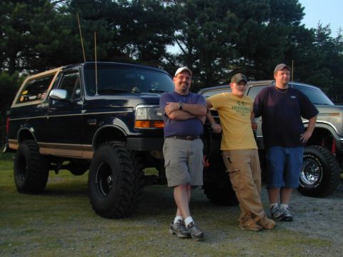 1988 Ford Bronco - Three men pose with a lifted dark blue Ford Bronco, likely from the 1990s, prominent off-road tires.