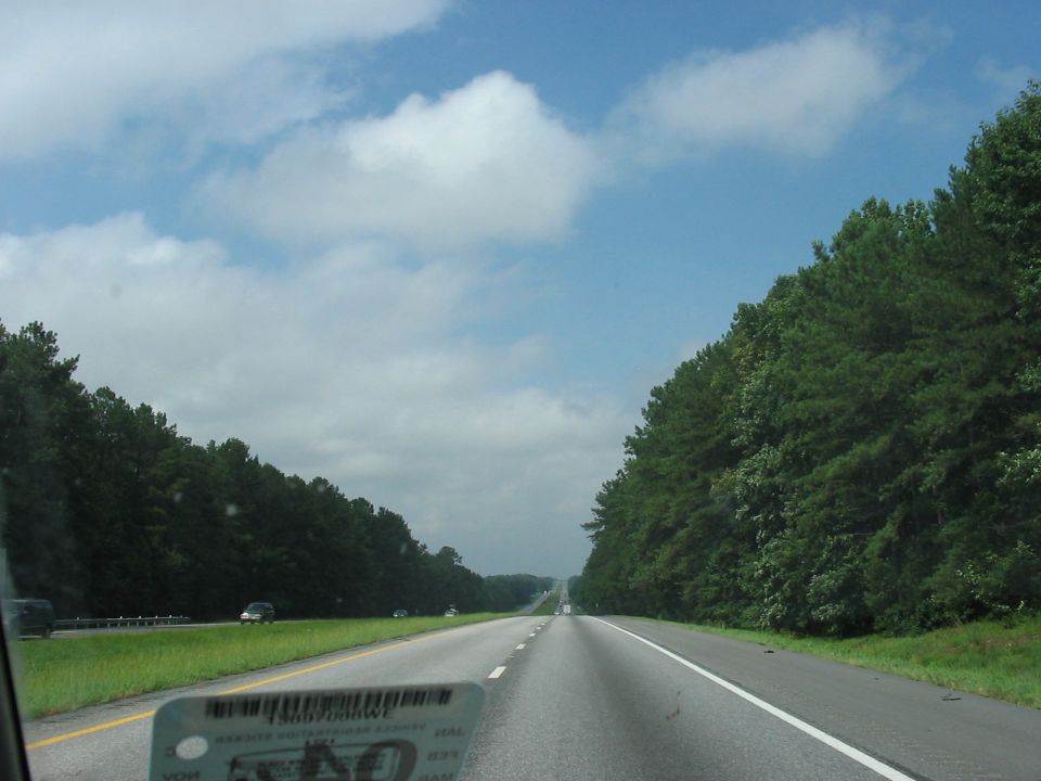 View from a car on a highway, looking ahead with trees lining both sides and a cloudy sky