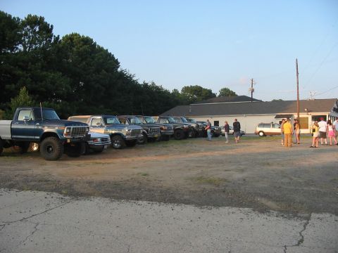1988 Ford Bronco - Several vintage Ford trucks and SUVs are parked in a lot near a building on a clear day.