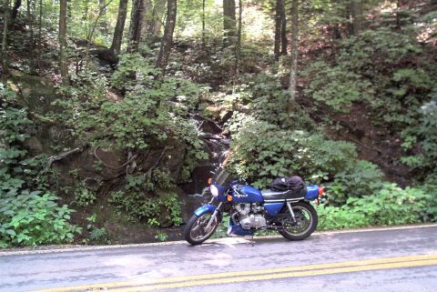 1985 Ford LTD Crown Victoria - A blue motorcycle is parked on the side of a road with a wooded area and a waterfall in the background.