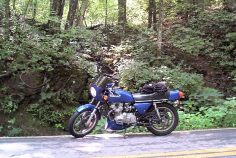 1985 Ford LTD Crown Victoria - A blue Suzuki motorcycle is parked on the side of a road with trees and foliage in the background.
