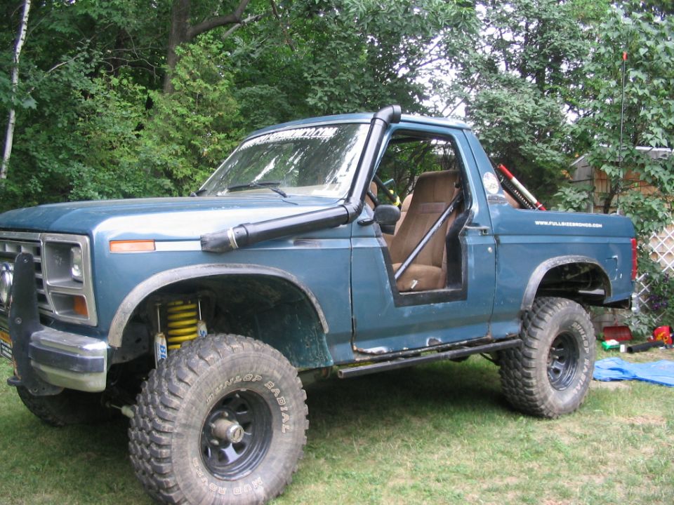 Blue 1980s Ford Bronco with doors and top removed, lifted on mud tires in a yard