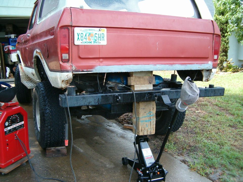 Red and white 1978 Ford Bronco rear end raised on blocks and jack, with a welding machine nearby