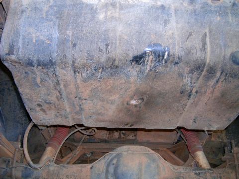 1978 Ford Bronco - Undercarriage view looking up at a dirt-caked steel fuel tank with a fresh weld repair and sender wiring on its face, mounted above the rea…