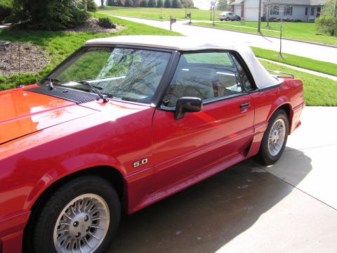 1990 Ford Mustang - A red convertible with a white top is parked on a driveway, showing its side profile and a '5.0' badge.