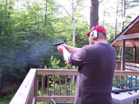 2001 Ford Crown Victoria - A man wearing a red cap and ear protection fires a handgun on a wooden deck, with trees and a cabin in the background.