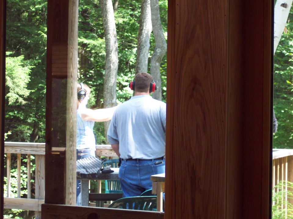 Two people wearing hearing protection stand on a deck, one aiming a rifle