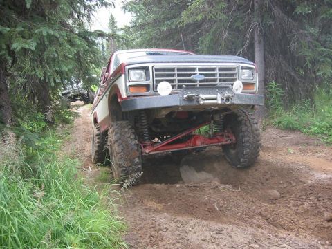 1985 Ford Bronco - A lifted 1985 Ford Bronco navigates a muddy, wooded trail, with another vehicle visible in the background.