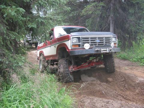 1985 Ford Bronco - Two off-road vehicles navigate a challenging dirt path through a wooded area.