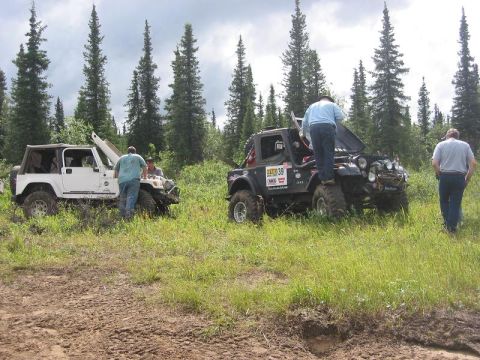 1985 Ford Bronco - Two off-road vehicles are stopped in a grassy field, with people attending to them under a cloudy sky.