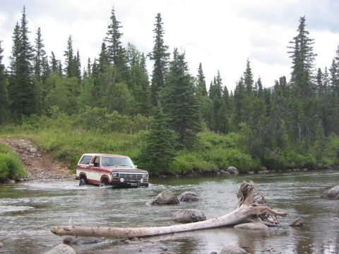 1985 Ford Bronco - A red and white SUV navigates a rocky riverbed surrounded by dense forest.