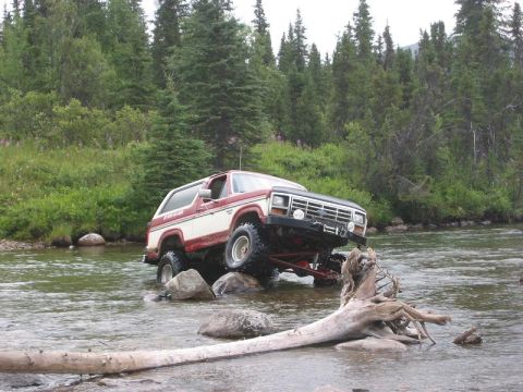 1985 Ford Bronco - Ford Bronco crosses a shallow river, balancing on rocks with a fallen tree nearby.