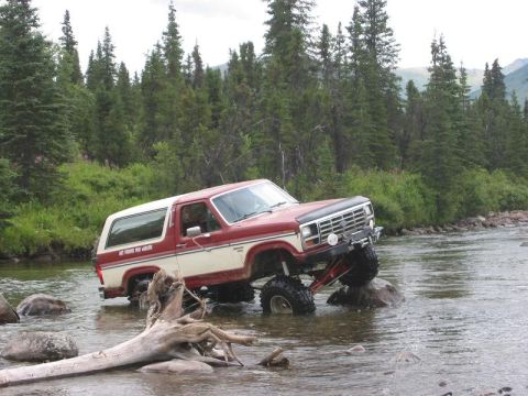 1985 Ford Bronco - A lifted Ford Bronco navigates a shallow riverbed, its tires gripping rocks amidst a backdrop of dense pine forest.