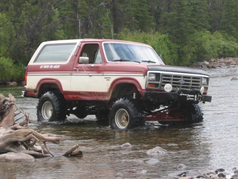 1985 Ford Bronco - This lifted Ford Bronco navigates a rocky riverbed surrounded by trees.