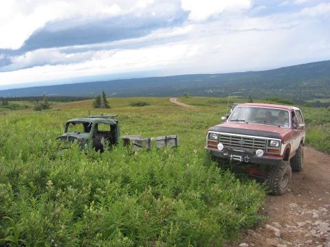 1985 Ford Bronco - A red and white 1985 Ford Bronco is parked on a dirt road in a scenic, hilly landscape, with an abandoned green truck visible in the tall g…