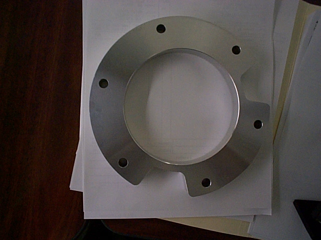Machined aluminum adapter plate with six bolt holes resting on a desk