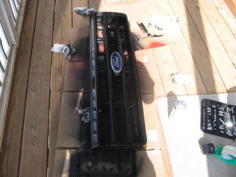 1988 Ford Bronco - A black Ford grille, likely from a 1988 Bronco, is laid out on a wooden deck for painting.