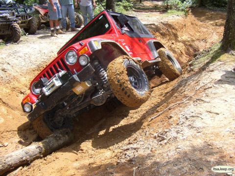 1984 Ford Bronco - Red Jeep Wrangler struggling on a steep, muddy trail with a log obstacle.