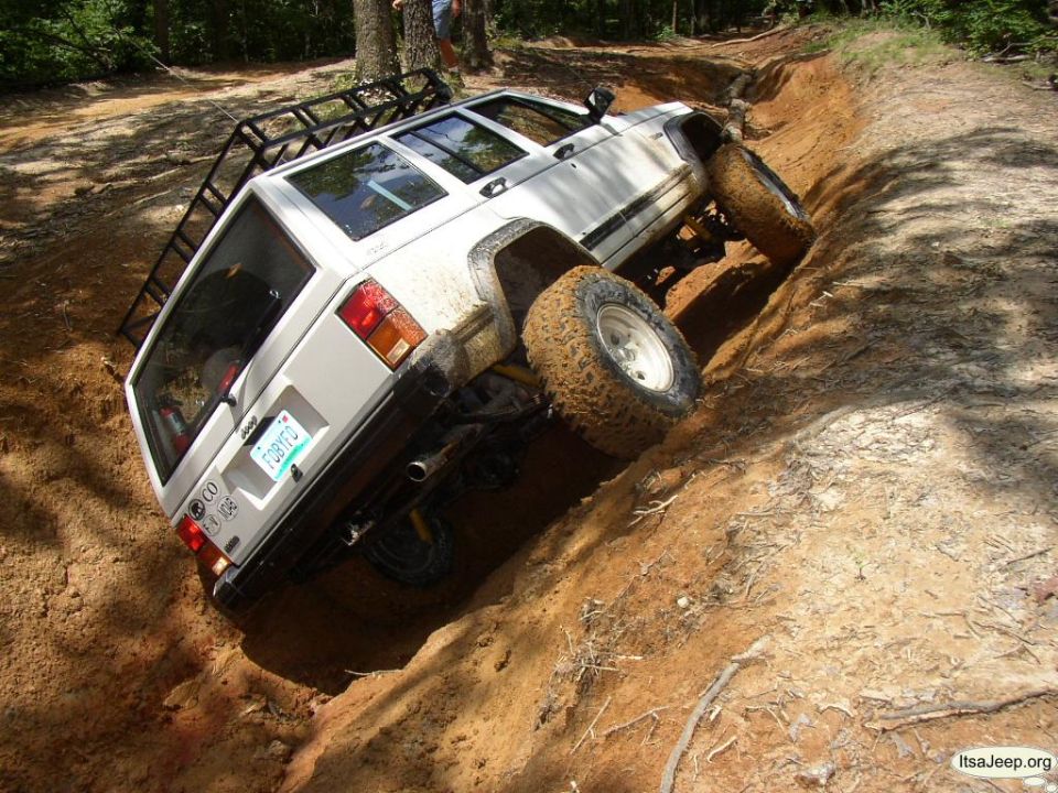 White Jeep Cherokee XJ with off-road tires climbing a dirt embankment, roof rack visible