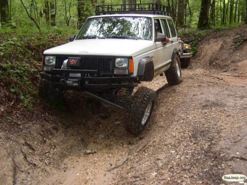 1984 Ford Bronco - A white Jeep Cherokee, modified for off-roading, tackles a challenging dirt path with another vehicle visible behind it.