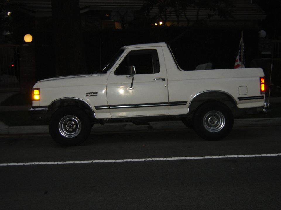 White Ford Bronco with hardtop removed parked on a street at night, American flag at the rear