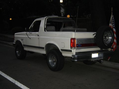 1988 Ford Bronco - This white Ford Bronco is parked on a street at night, showing its rear profile and spare tire.