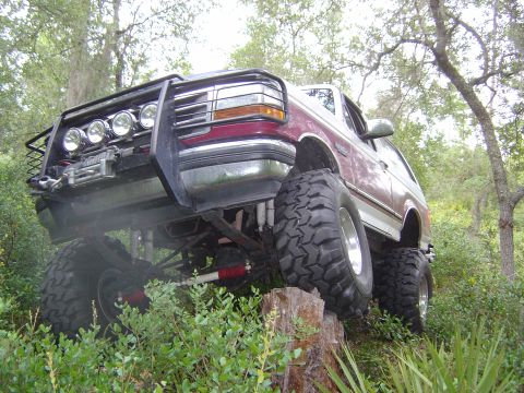 1993 Ford Bronco - Modified Ford Bronco with extreme suspension and tires navigates an obstacle in a wooded area.