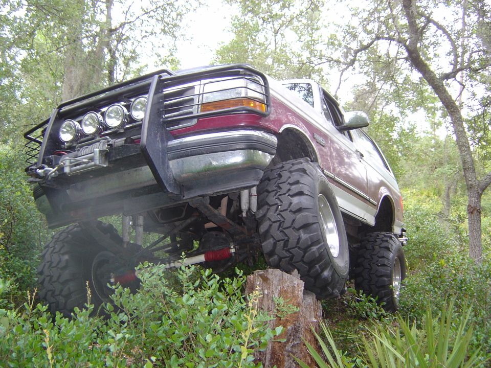 Maroon and silver lifted Ford Bronco with large off-road tires climbing a tree stump