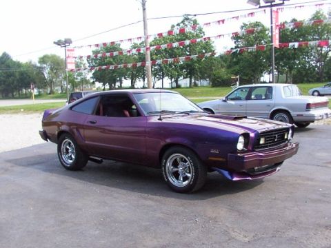 1978 Ford Mustang II - A purple 1977 Ford Mustang II coupe is parked in front of a dealership with another car visible behind it.