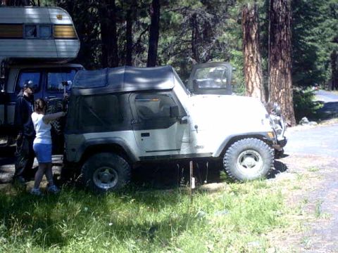 1995 Ford Bronco - A white Jeep Wrangler with a soft top is parked in a wooded area next to a camper trailer.