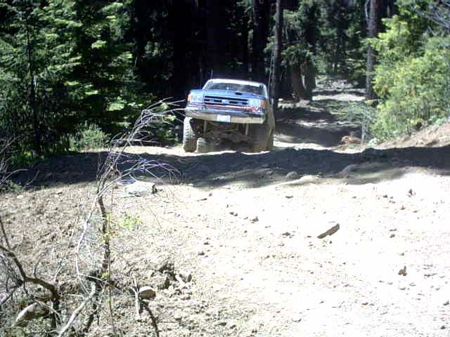 Lifted light blue Ford on large tires climbing a dusty forest dirt trail
