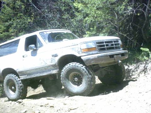 1995 Ford Bronco - This lifted Ford Bronco is navigating a dusty, off-road trail with trees in the background.
