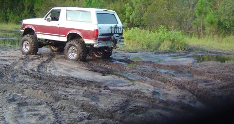 1993 Ford Bronco - Rear three-quarter view of a lifted burgundy and white full-size Bronco flexing over rutted mud with a driver at the wheel, one front tire…