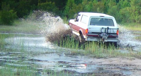 1993 Ford Bronco - Three-quarter rear view of a white-topped maroon Ford Bronco charging through a flooded grassy mud flat, kicking a tall rooster tail of wat…