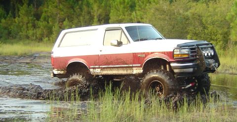 1993 Ford Bronco - Full side profile of a lifted burgundy and white full-size Bronco crawling through shallow mud and grass, with the BRONCO fender badge visi…