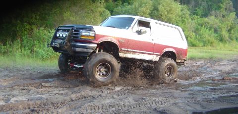 1993 Ford Bronco - Side-on action view of a lifted burgundy and white full-size Bronco climbing out of a mud pit with the front end raised and mud spraying of…
