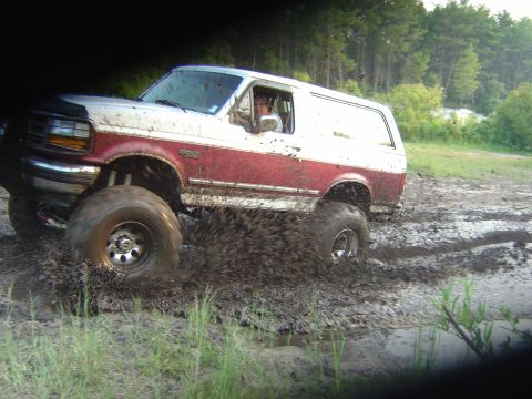 1993 Ford Bronco - Side view of a lifted burgundy and white full-size Bronco powering through a mud pit with the driver leaning out the open window and mud fl…