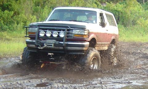 1993 Ford Bronco - Front three-quarter action shot of a burgundy and white full-size Bronco churning through a mud pit with the driver visible through the win…