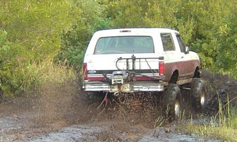 1993 Ford Bronco - Rear view of a burgundy and white full-size Bronco driving away through a muddy trail, spraying mud from the tires. The tailgate carries a…