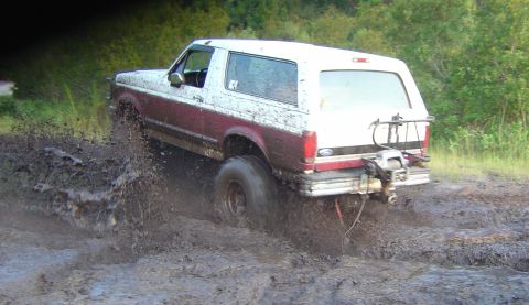 1993 Ford Bronco - Rear three-quarter action view of a mud-covered burgundy and white full-size Bronco driving away through a churned mud pit, slinging mud of…