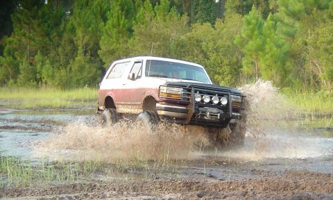 1993 Ford Bronco - Action shot of a two-tone burgundy and white full-size Bronco blasting through a shallow mud pit, throwing a wide spray of muddy water off…