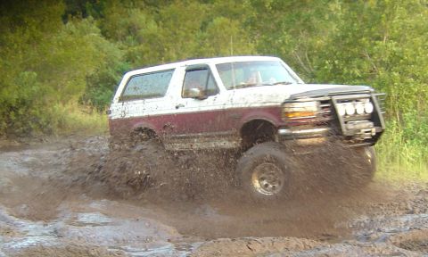 1993 Ford Bronco - Action side shot of a lifted white-over-maroon two-door Ford Bronco blasting through a mud pit, spraying muck off its mud-terrain tires and…