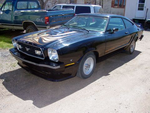 1978 Ford Mustang II - Black Ford Mustang II hatchback with running-horse grille emblem and hood decal, parked on a dirt drive near a pickup.