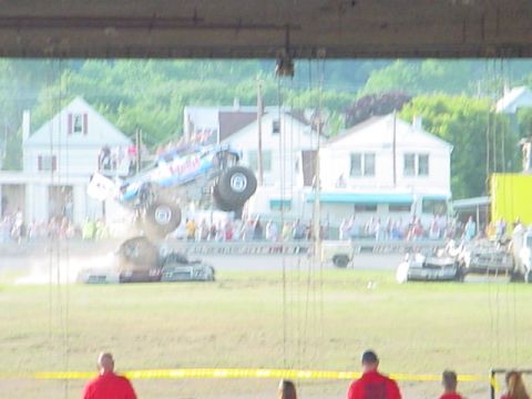 1979 Ford Bronco - A monster truck performs a jump over several cars in a field with spectators in the background.
