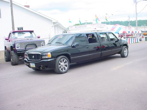 1979 Ford Bronco - A very long black pickup truck limo is parked next to a lifted purple pickup truck at an outdoor event.
