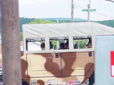 1979 Ford Bronco - A camouflaged truck with a canvas top carries passengers on a road with East and West street signs visible.