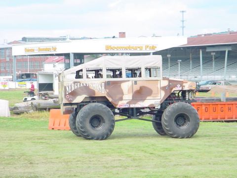 1979 Ford Bronco - A camouflage-painted monster truck, labeled 'SERGEANT SMASH', sits on a grassy field with a fairground in the background.