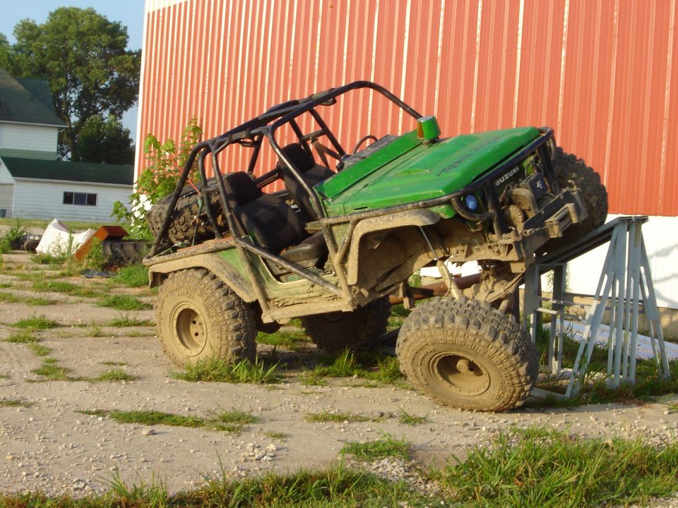 Green off-road buggy with roll cage, lifted on metal stands in front of a red barn