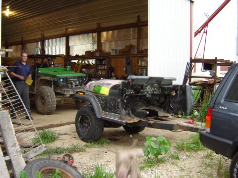 1992 Ford Bronco - stripped vehicle chassis under a metal shed roof, green side-by-side and dog nearby in a workyard.